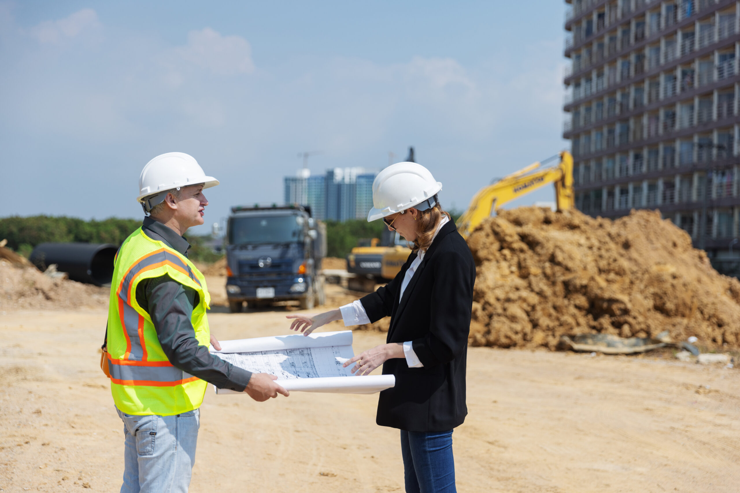 Two construction professionals discuss architectural blueprints at a building site, surrounded by dirt piles, a dump truck, and an excavator, highlighting urban development assessment rules and project coordination.
